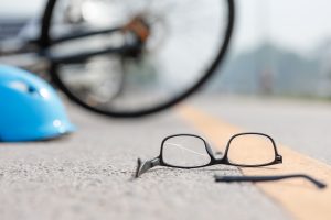 A pair of upside-down broken glasses on a road with a blue helmet and the wheel of a bike also on the road in the background. 