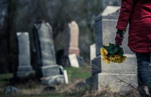 A person in a red coat stands in front of a gravestone and holds a bouquet of yellow sunflowers.