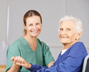 A young female care worker wearing a green uniform holding hands and sat opposite an elderly woman in a wheelchair wearing a purple jumper. Both are smiling. 