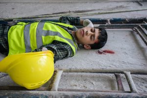 A male construction worker lying unconscious on the ground with some blood by his head.