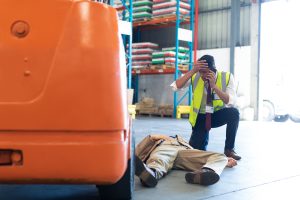 A forklift truck driver laying on the ground with serious injuries while a colleague kneeling over him phones for help