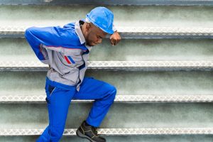 A worker in a blue helmet with blue overalls leans against a stairwell after suffering a back injury