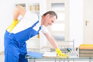 A man in blue overalls and yellow rubber gloves leans over a table in pain